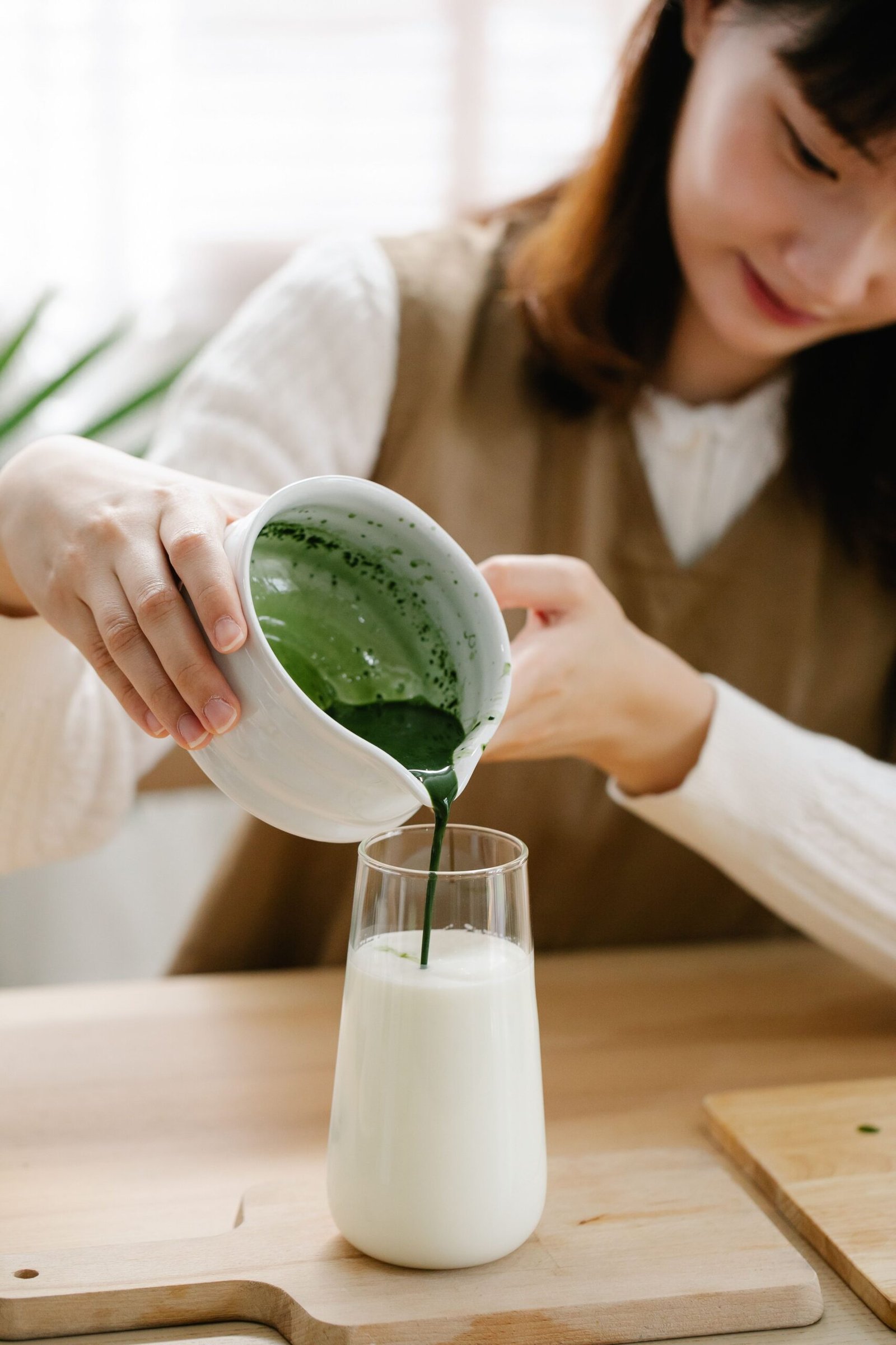 A woman pouring matcha into a glass of milk on a wooden surface indoors.